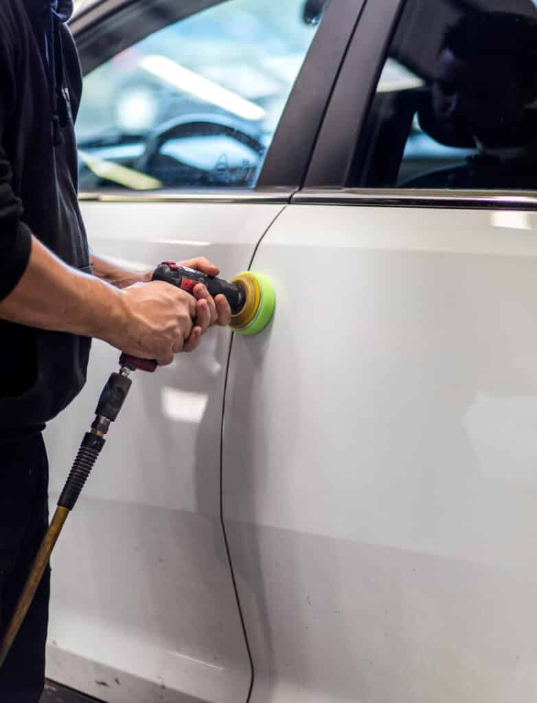 Homme en sweat à capuche noir polissant une porte de voiture blanche avec une polisseuse pneumatique et un tampon vert-jaune.