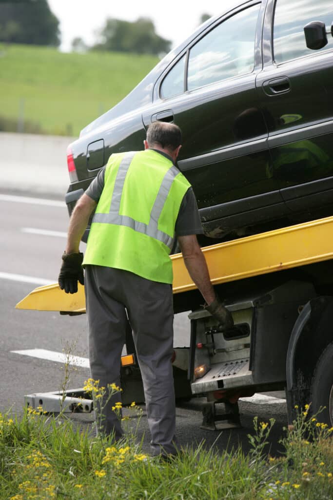 Ouvrier avec gilet haute visibilité chargeant une voiture noire sur dépanneuse jaune au bord de la route.
