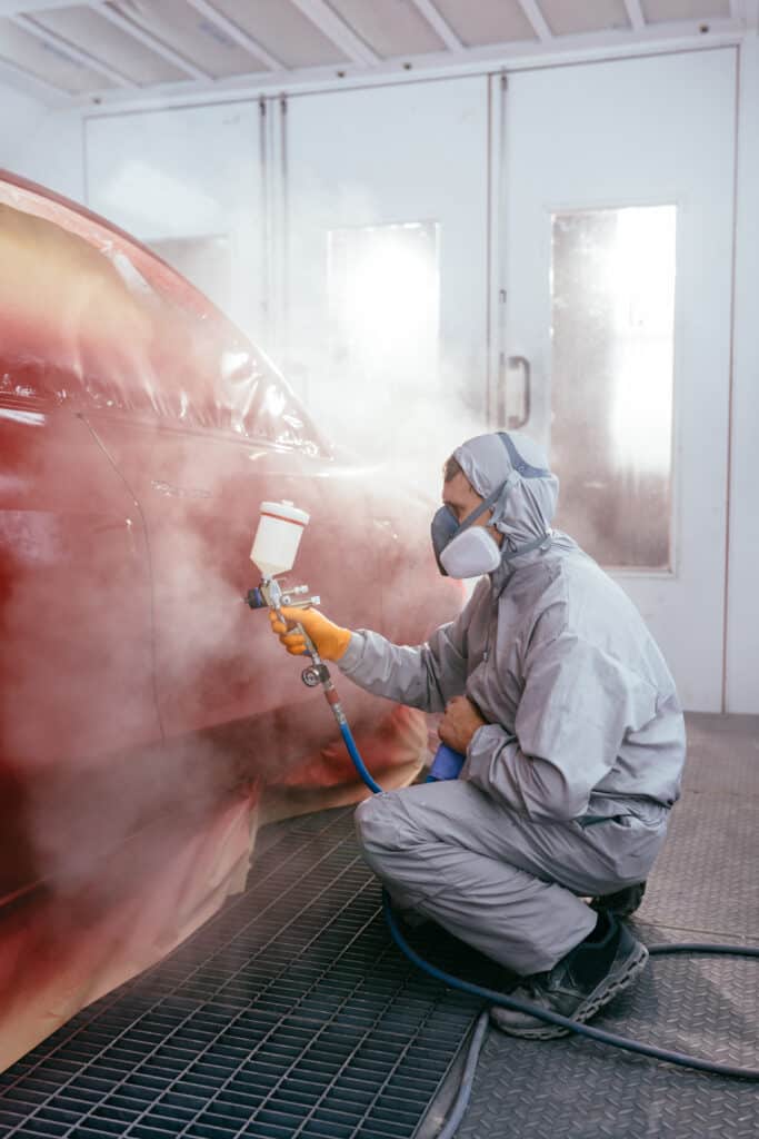 Carrossier en combinaison de protection et masque, pulvérisant de la peinture rouge sur une voiture dans une cabine de peinture.