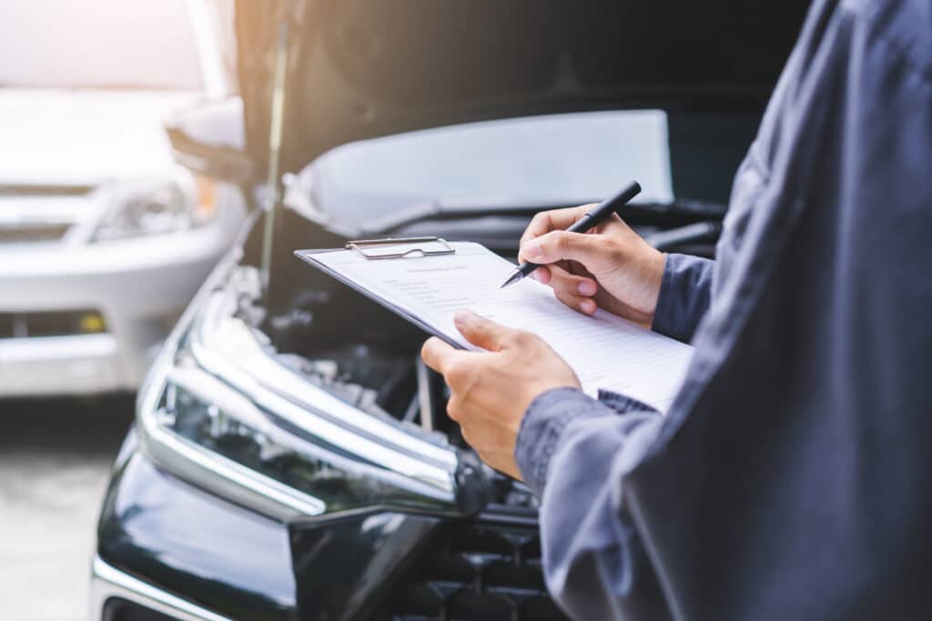 Mécanicien en uniforme remplissant un formulaire sur un presse-papiers devant un capot de voiture ouvert, inspection automobile.