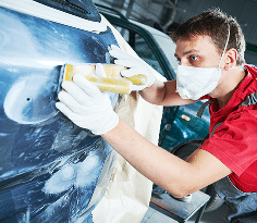 Carrossier masqué ponce avec une cale un panneau de voiture bleu pour préparation peinture, portant gants blancs.