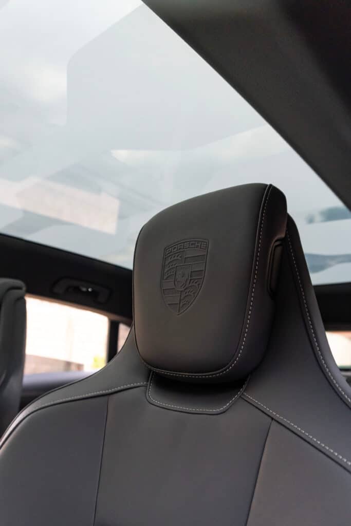 Intérieur Porsche Cuir : Blason Appui-Tête et Toit Ouvrant Gros plan sur l'appui-tête en cuir noir d'un siège Porsche, avec le blason gaufré et des surpiqûres blanches, sous un toit ouvrant vitré.